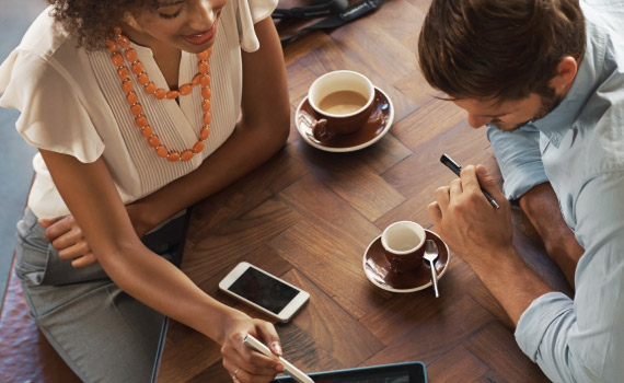Two adults appear to be looking at a tablet while enjoying coffee.