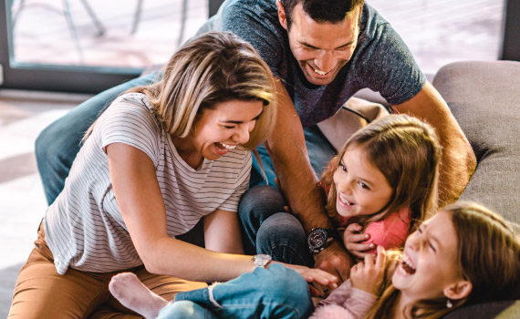 A happy mother, father and two daughters pile on a couch in laughter.